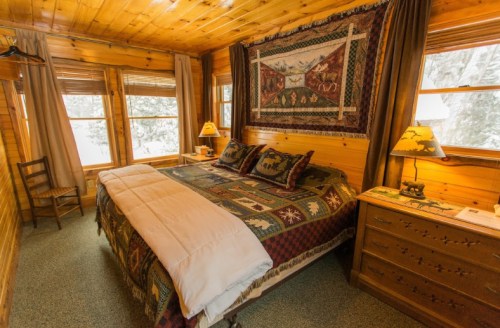Cozy wooden bedroom with a quilted bedspread, two lamps, and large windows showing a snowy landscape outside.