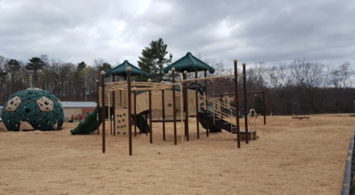 Playground with wooden structures, slides, and a climbing dome, surrounded by tan mulch and overcast skies.