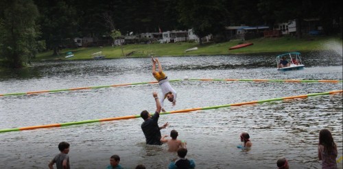 A child flips in the air above a lake while being caught by an adult, surrounded by other kids enjoying the water.