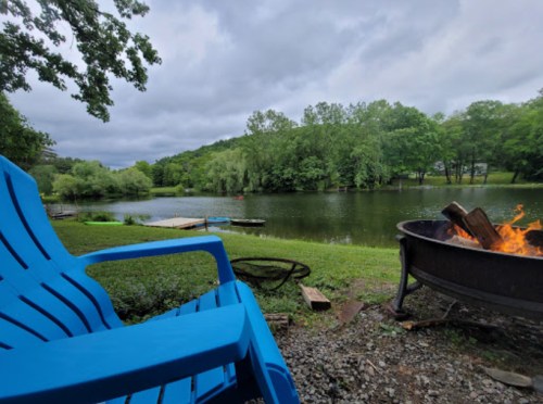 A blue chair overlooks a calm lake surrounded by trees, with a fire pit in the foreground and cloudy skies above.