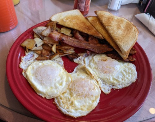 A red plate with two fried eggs, crispy bacon, toast, and seasoned potato cubes.