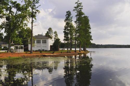 A serene lakeside view featuring a white house, tall trees, and calm water reflecting the sky.