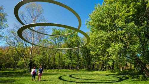 Two people stand in a green park beneath large, suspended circular sculptures against a clear blue sky.
