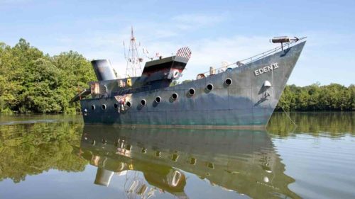 A weathered ship named "EDEN II" anchored in calm waters, surrounded by lush greenery and a clear blue sky.