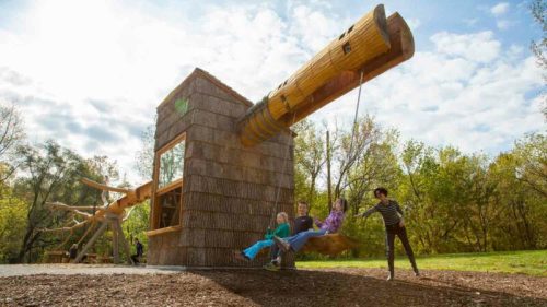A unique playground structure resembling a treehouse with a large wooden cannon and children playing on swings.