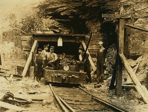Historic black-and-white photo of miners near a coal mine entrance, with a small cart on tracks and tools scattered around.