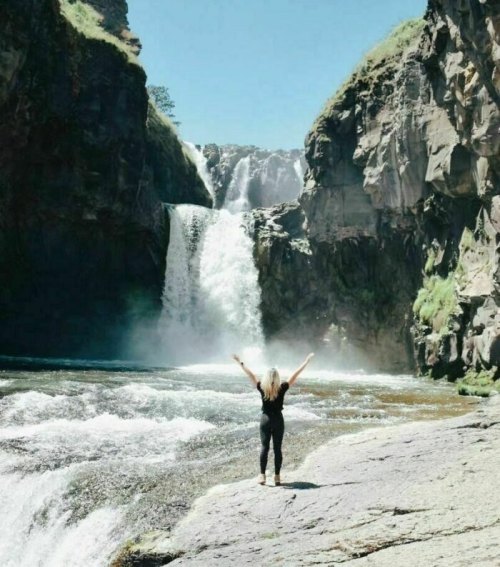 A person stands with arms raised in front of a majestic waterfall, surrounded by rocky cliffs and lush greenery.