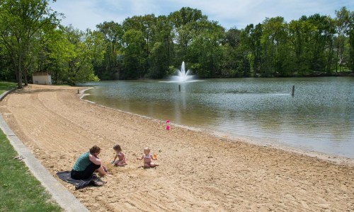 A woman and two children play on a sandy beach by a calm lake with a fountain and trees in the background.