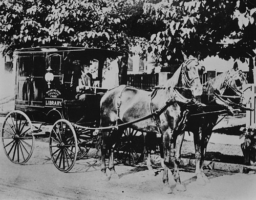 A historic black-and-white photo of a horse-drawn library cart with a driver and two horses.