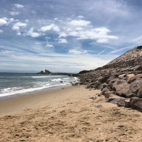 A sandy beach with rocky shorelines, gentle waves, and a cloudy blue sky in the background.