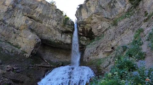 A tall waterfall cascades down rocky cliffs, surrounded by lush greenery and rugged terrain.