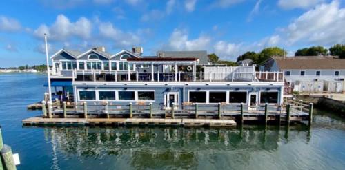 A waterfront building with a blue exterior, featuring outdoor seating and a dock, under a partly cloudy sky.