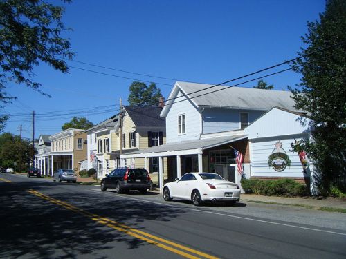 A quiet street lined with charming houses and parked cars under a clear blue sky. American flags are displayed.