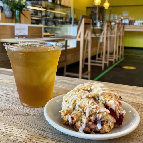 A scone drizzled with icing on a plate, next to a cup of iced tea, with a café interior in the background.