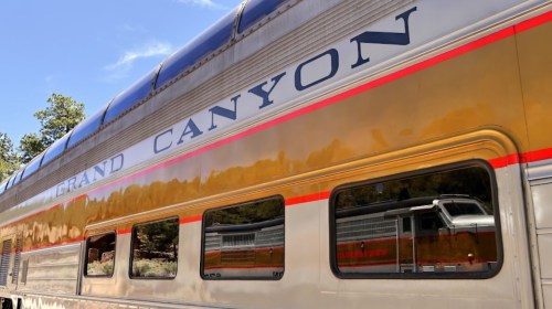 A close-up of a train car with "Grand Canyon" written on the side, reflecting trees and scenery in its windows.