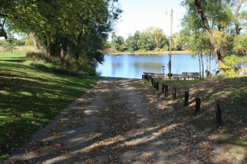 A serene path lined with trees leads to a calm lake, surrounded by greenery and scattered autumn leaves.