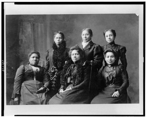 A historic black-and-white photo of six women in 19th-century attire, posing together in a studio setting.