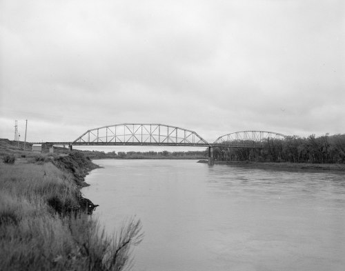 A black and white photo of a bridge spanning a river, with overcast skies and trees along the riverbank.