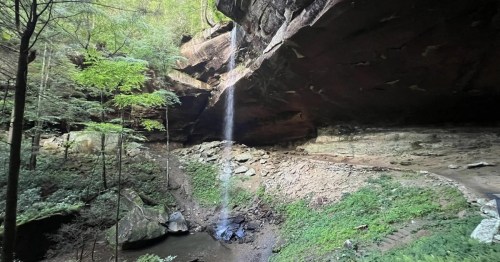 A serene waterfall cascades from a rocky overhang into a lush, green valley surrounded by trees.