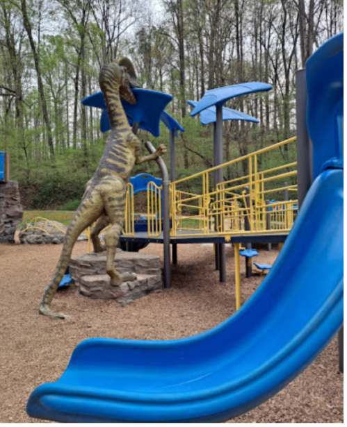 A dinosaur sculpture interacts with a playground featuring blue slides and yellow structures, surrounded by trees.