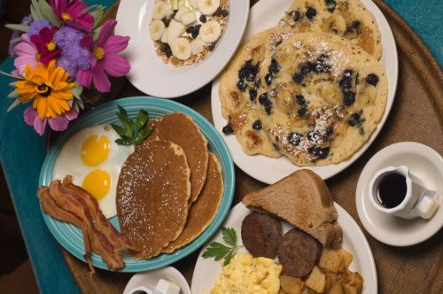 A colorful breakfast spread featuring pancakes, eggs, bacon, and fresh fruit, surrounded by vibrant flowers.
