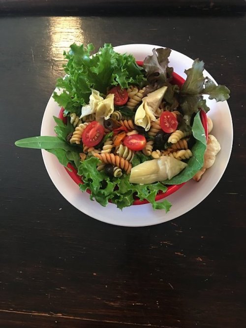 A colorful salad with pasta, cherry tomatoes, artichokes, and mixed greens in a red bowl on a dark surface.