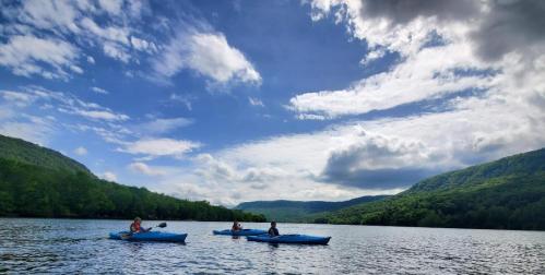 Three kayakers paddle on a calm lake surrounded by lush green hills and a partly cloudy sky.