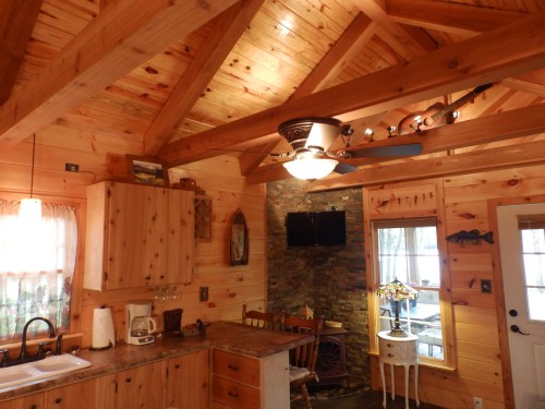 Cozy wooden cabin interior featuring a kitchen, dining area, and stone fireplace with a ceiling fan and natural light.
