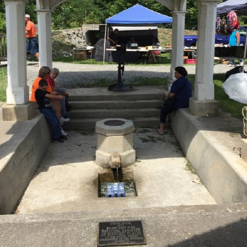 A small water fountain under a gazebo, with people sitting nearby and tents in the background.