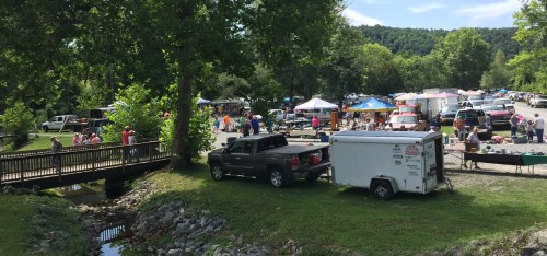 A bustling outdoor market with tents, vendors, and people shopping, surrounded by trees and a small creek.