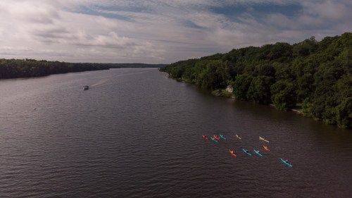 Aerial view of a river with kayakers in colorful boats and a small boat in the distance, surrounded by lush greenery.