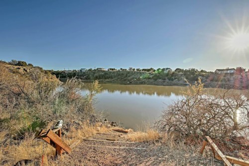 A serene river scene with sun shining, surrounded by dry vegetation and distant houses under a clear blue sky.