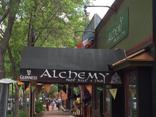 Sign for "Alchemy" pub with a Guinness logo, surrounded by colorful banners and trees lining the street.