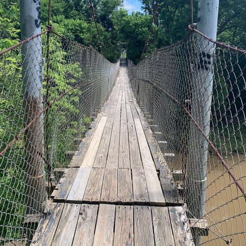 A long, narrow suspension bridge made of wooden planks, surrounded by greenery and spanning a river below.