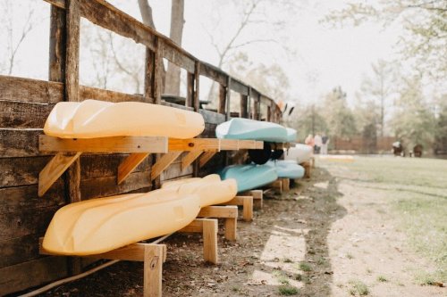 Colorful kayaks are neatly stacked on a wooden rack beside a grassy area in a sunny outdoor setting.