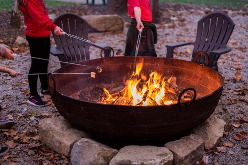 People roasting marshmallows over a large fire pit surrounded by chairs and autumn leaves.