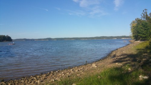A serene lakeside view with calm water, rocky shore, and green trees under a clear blue sky.