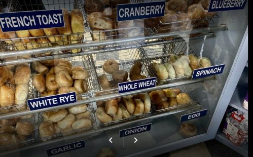 A display case filled with various bagels labeled: French Toast, Cranberry, Blueberry, White Rye, Whole Wheat, Spinach, Garlic, Onion, Egg.