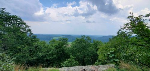 A scenic view of rolling hills and valleys under a cloudy sky, framed by lush green trees.