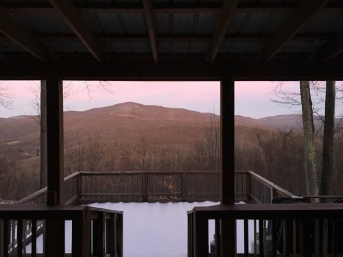 A serene view of mountains at dusk, framed by a wooden porch with snow on the ground.