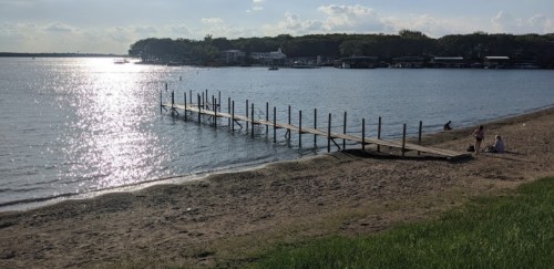 A wooden dock extends into a calm lake, with sandy beach and people sitting nearby under a sunny sky.