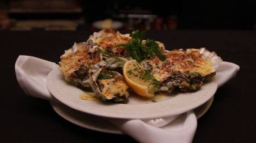 A plate of baked oysters topped with cheese, garnished with lemon and herbs, served on a white napkin.