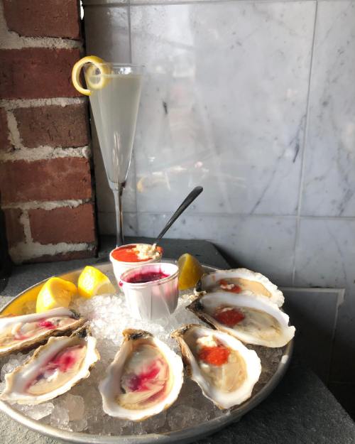 A platter of oysters on ice with lemon wedges, accompanied by a cocktail and dipping sauces.
