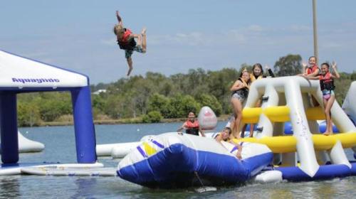 A child jumps off an inflatable structure into the water, while others play on nearby inflatable equipment.