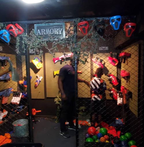 A dimly lit armory filled with colorful toy guns and balls, with two people inside exploring the equipment.