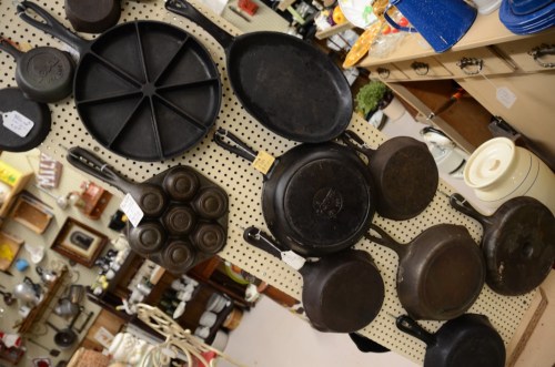 A collection of vintage cast iron cookware displayed on a pegboard in a shop, including skillets and a muffin pan.
