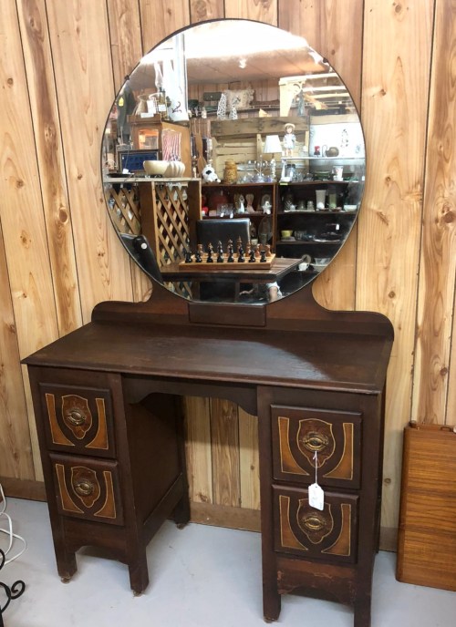 A vintage wooden dresser with a round mirror, featuring decorative drawer designs, set against a wooden wall.