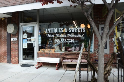 A storefront displaying "Antiques & Sweets" with a welcoming sign and various items visible through the window.