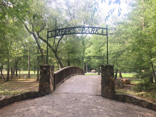 A stone bridge leads into Meeks Park, surrounded by lush green trees and a peaceful natural setting.