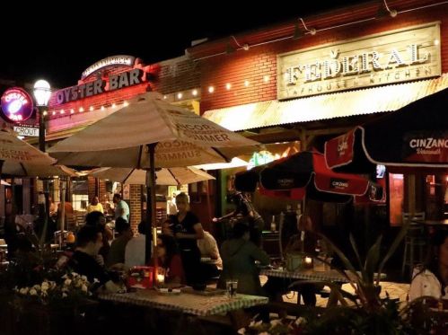 A lively outdoor dining scene at night, featuring illuminated restaurant signs and patrons enjoying their meals.